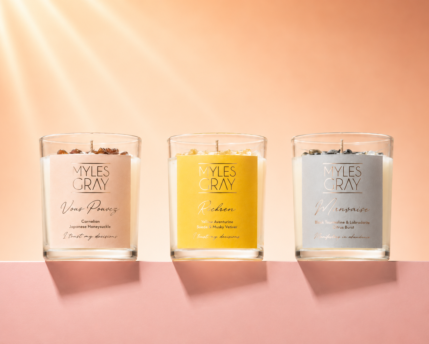 Three Myles Gray scented candles in clear glass jars are lined up on a pink surface against a peach background. The left candle has a pale pink label, the middle candle has a bright yellow label, and the right candle has a light blue-gray label.
